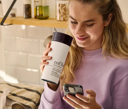 Kobieta trzyma kubek termiczny na kawę Coffee Dictionary i patrzy na telefon komórkowy.