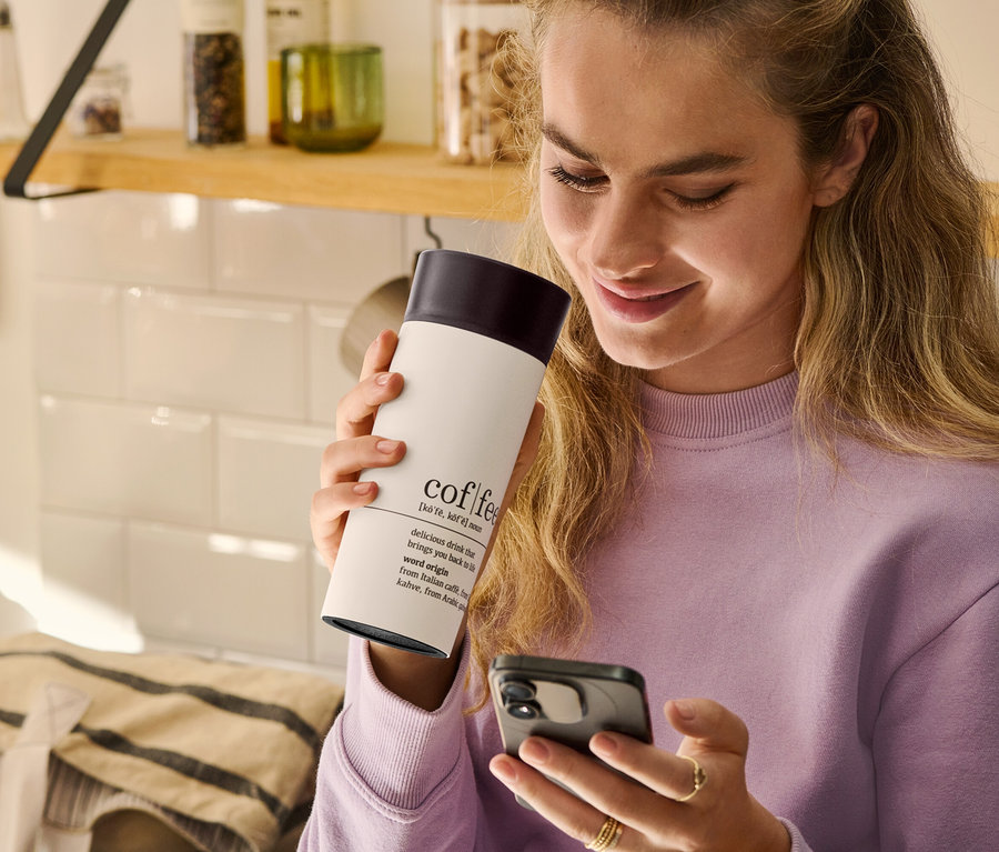 Kobieta trzyma kubek termiczny na kawę Coffee Dictionary i patrzy na telefon komórkowy.