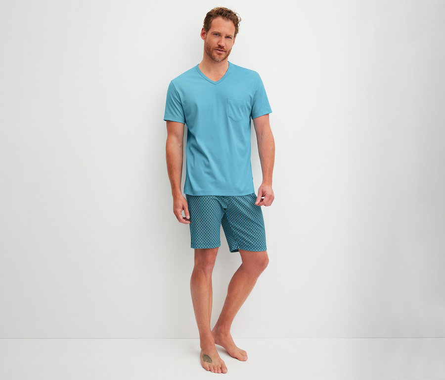 Mężczyzna stoi boso, ubrany w piżamę z krótkim rękawem i krótkimi spodenkami CALIDA Relax Streamline, składającą się z jasnoniebieskiego T-shirtu z dekoltem w serek i kieszenią oraz wzorzystych szortów.