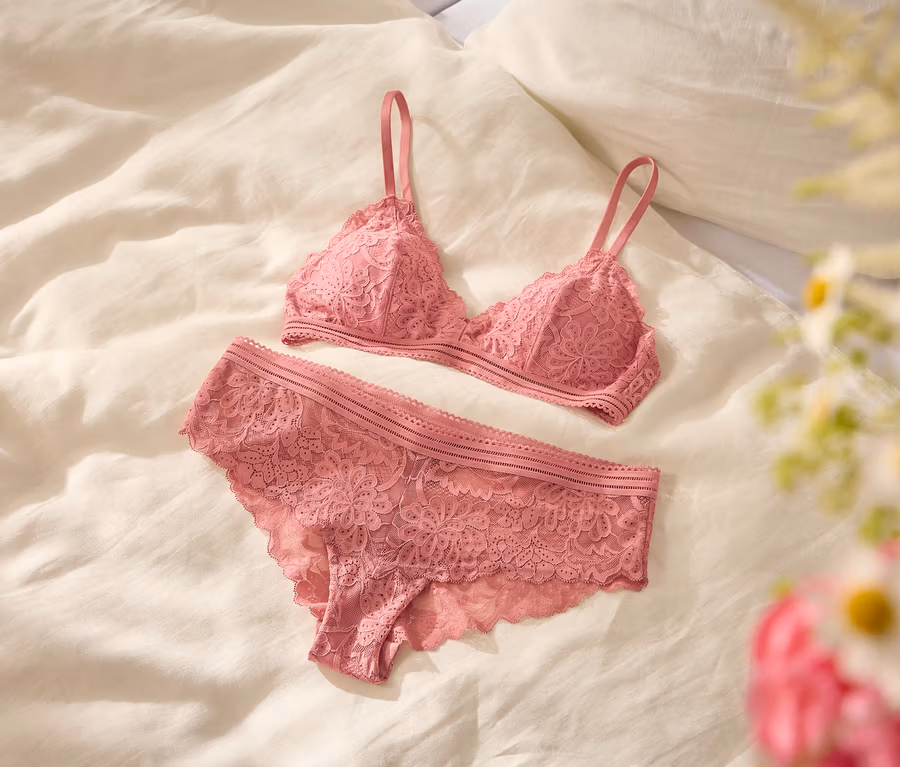 Różowy koronkowy biustonosz typu bralette i pasujące figi leżą płasko na białym prześcieradle, z rozmytym bukietem białych i różowych kwiatów na pierwszym planie po prawej.