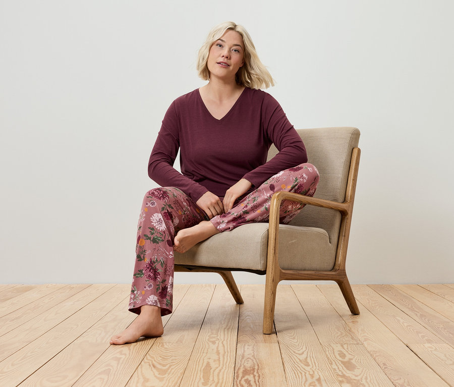 Blondwłosa kobieta siedzi w fotelu, ubrana w bordową koszulkę z długim rękawem i spodnie od piżamy w kwiatowy wzór.