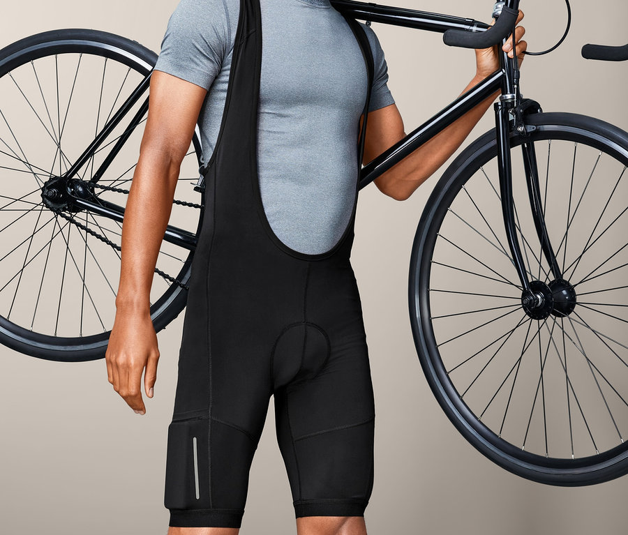 Mężczyzna w spodenkach rowerowych na szelkach niesie rower na ramieniu.