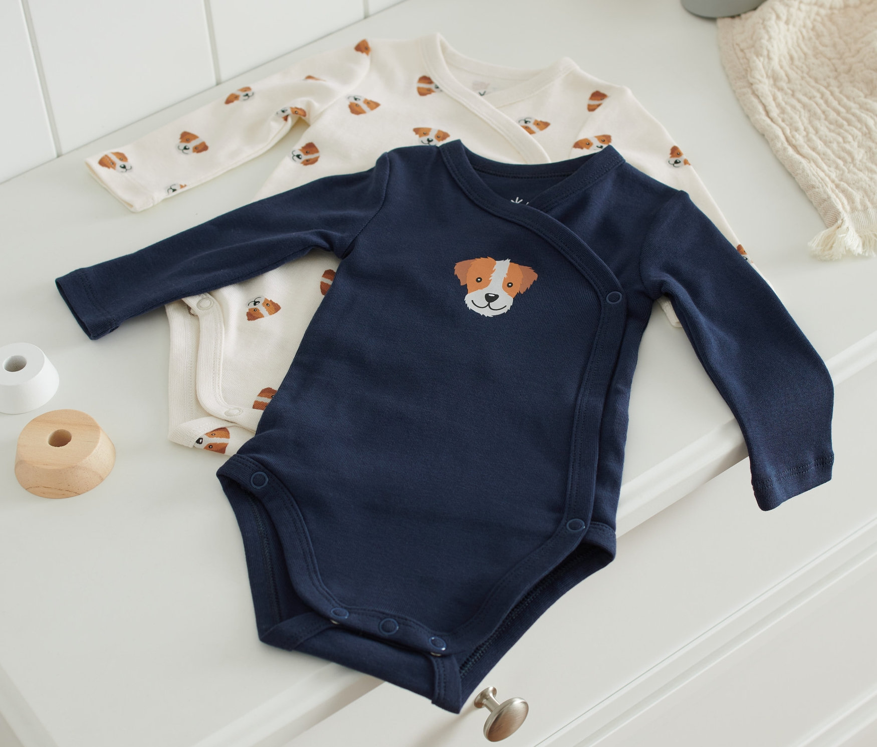 Dwa body leżą na białej komodzie. Jedno body jest granatowe z haftem przedstawiającym głowę psa, drugie jest kremowe z nadrukiem psów.