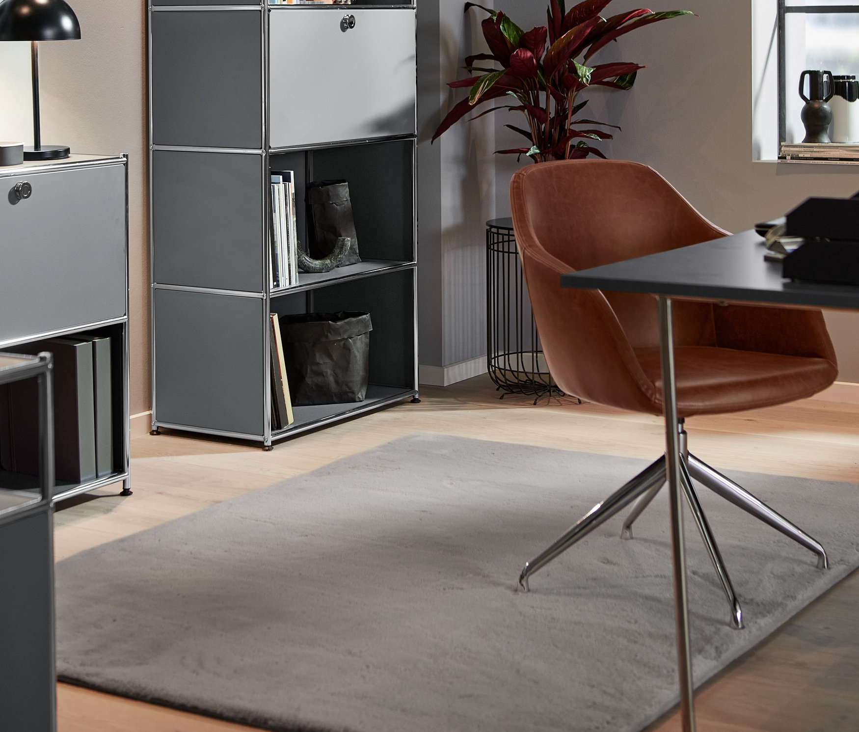 W pokoju stoi obrotowy fotel ze sztucznej skóry z wyściełanym siedziskiem, wysoka, metalowa szafka z przegrodą z klapą, montowaną w dowolnym miejscu i metalowa lampa stołowa LED. Na podłodze leży szary dywan z długim włosiem.