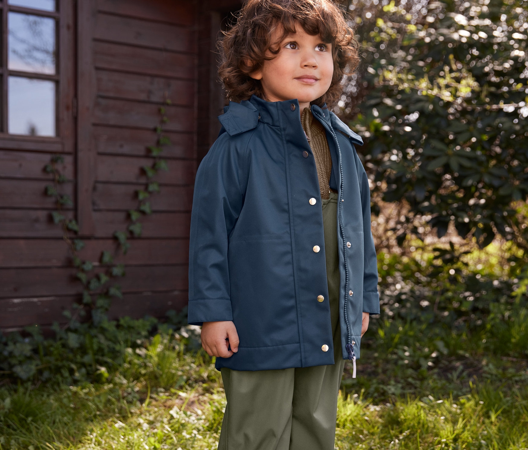 Chłopiec stoi na trawie na zewnątrz, ubrany w kalosze, spodnie przeciwdeszczowe i kurtkę przeciwdeszczową.