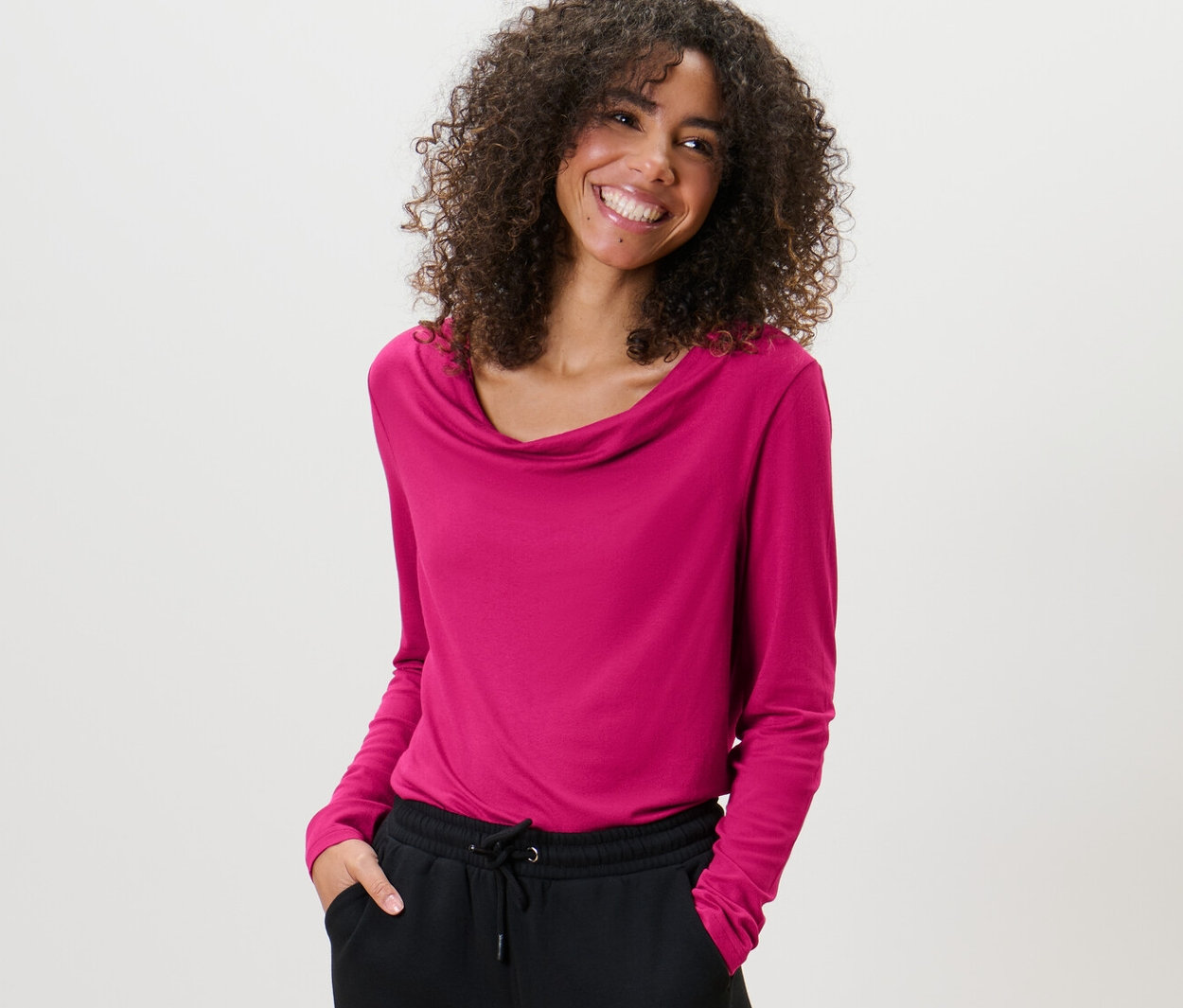 Kobieta z kręconymi włosami ma na sobie bluzkę z lejącym dekoltem i czarne spodnie.