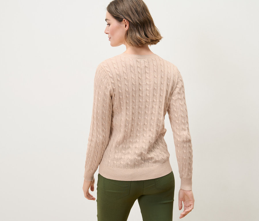 Kobieta z brązowymi włosami pozuje tyłem do kamery, ubrana w kremowy sweter z warkoczowym wzorem.