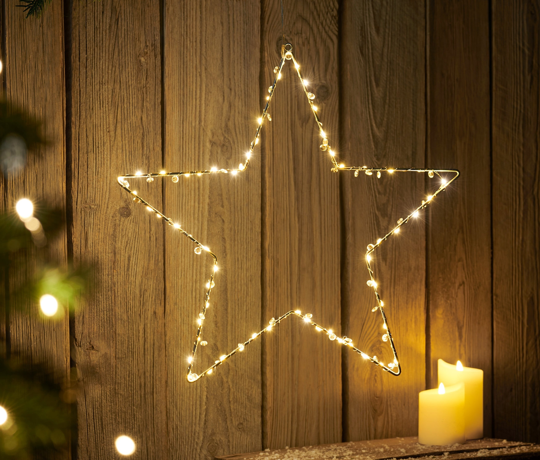 Duża zewnętrzna gwiazda LED wisi na drewnianej ścianie obok dwóch białych świec.