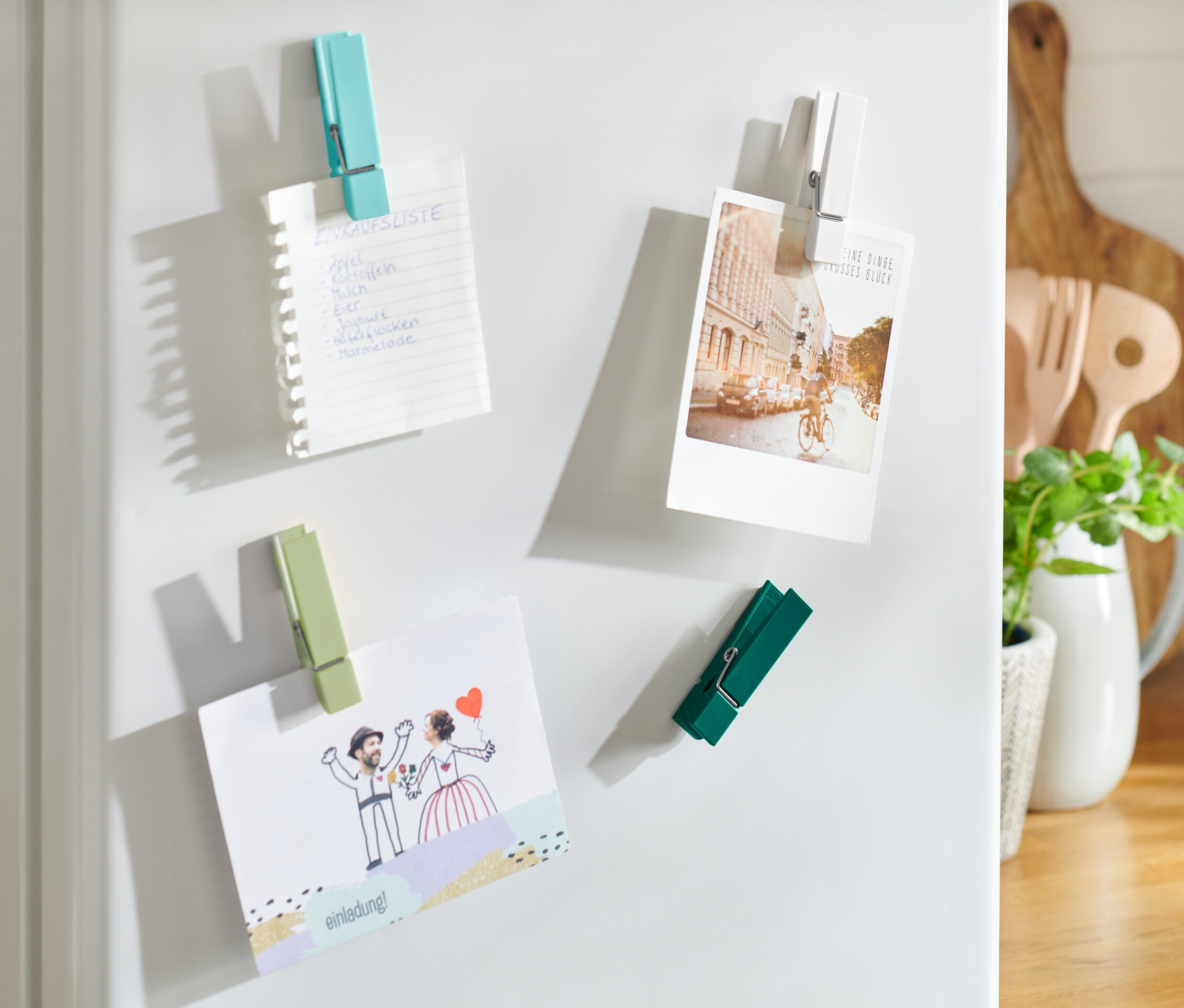 Cztery magnesy na lodówkę trzymają listę zakupów, pocztówkę i zaproszenie.
