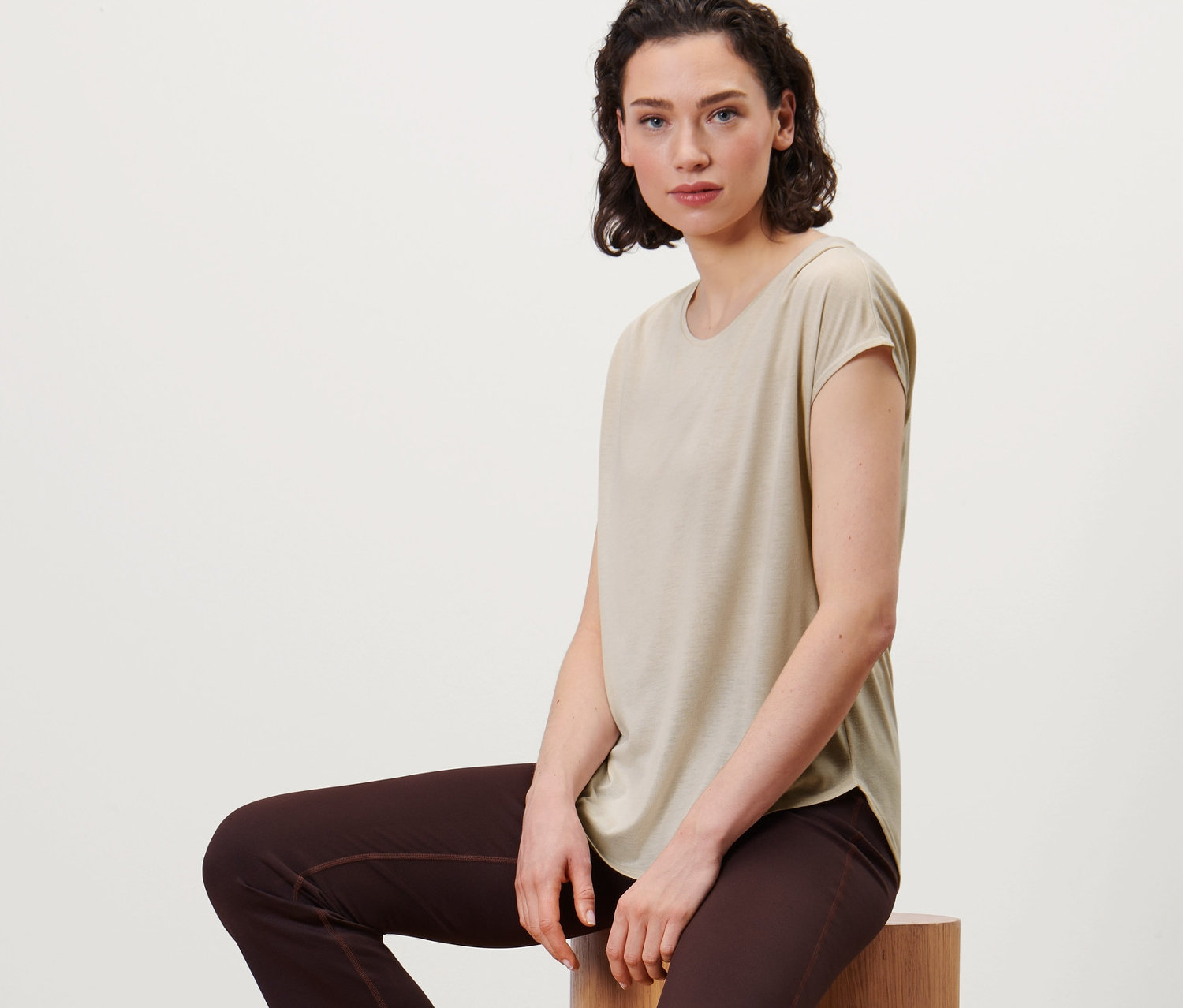 Kobieta siedzi na drewnianych cylindrach, ubrana w beżową koszulkę sportową i brązowe spodnie.