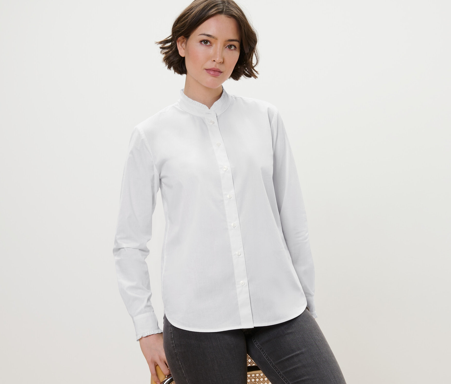 Kobieta pozuje na krześle, ubrana w białą bluzkę z falbankami i szare dżinsy.