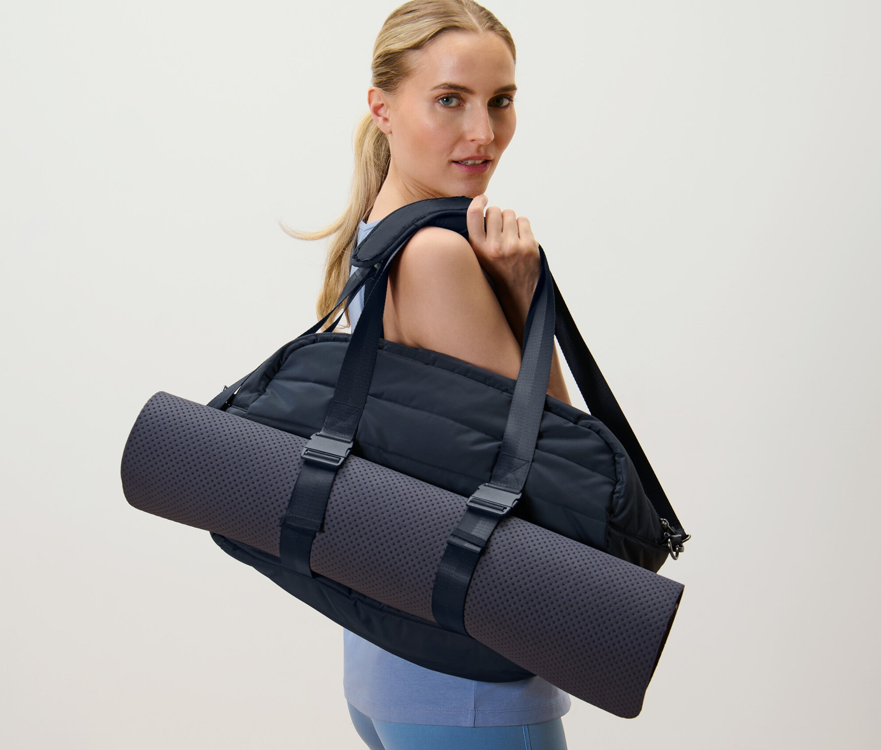 Blondynka niesie czarną torbę sportową i matę do jogi na ramieniu.