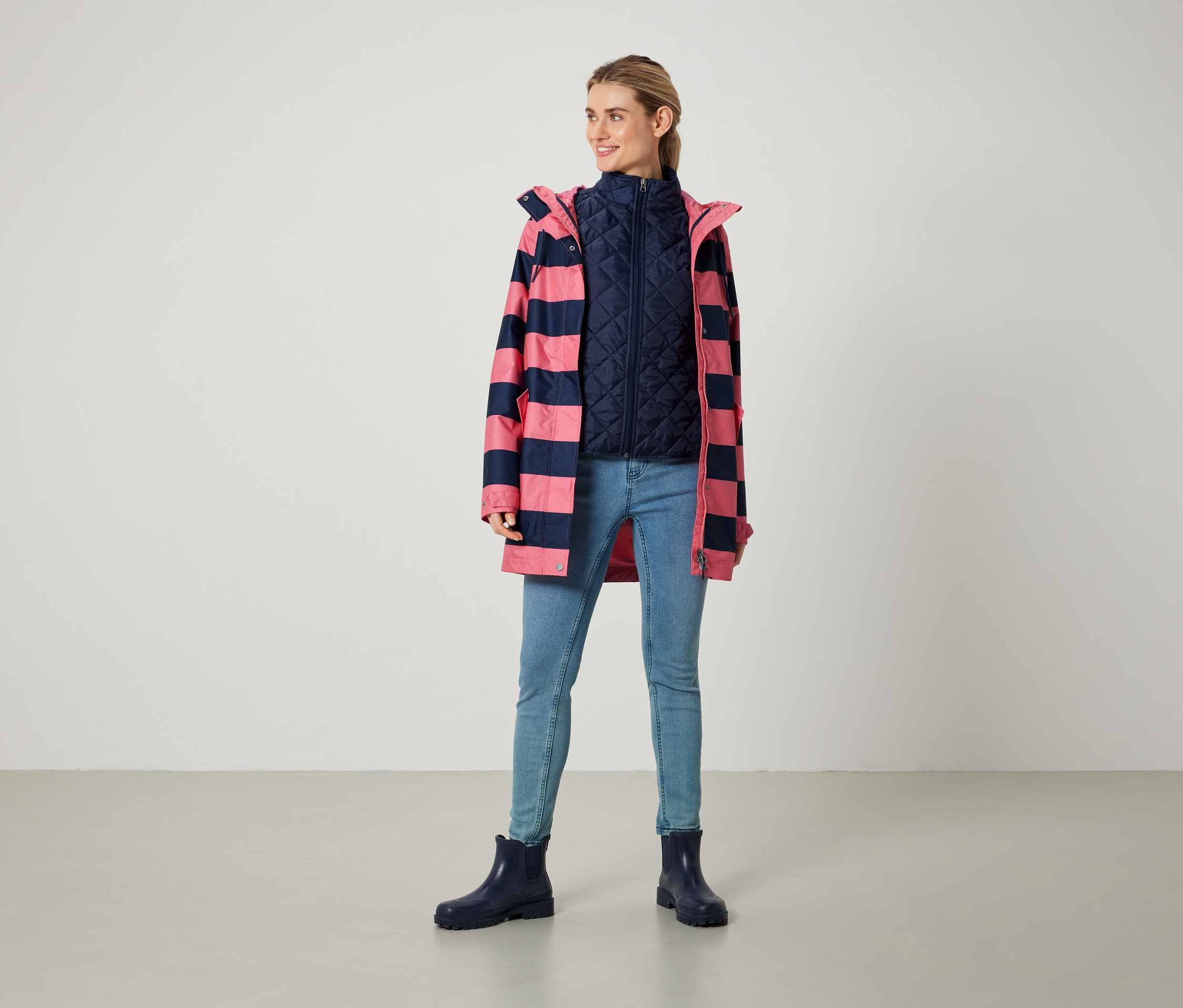 Kobieta stoi, ubrana w różowo-granatowy płaszcz w paski, granatową kamizelkę, dżinsy i niebieskie buty.