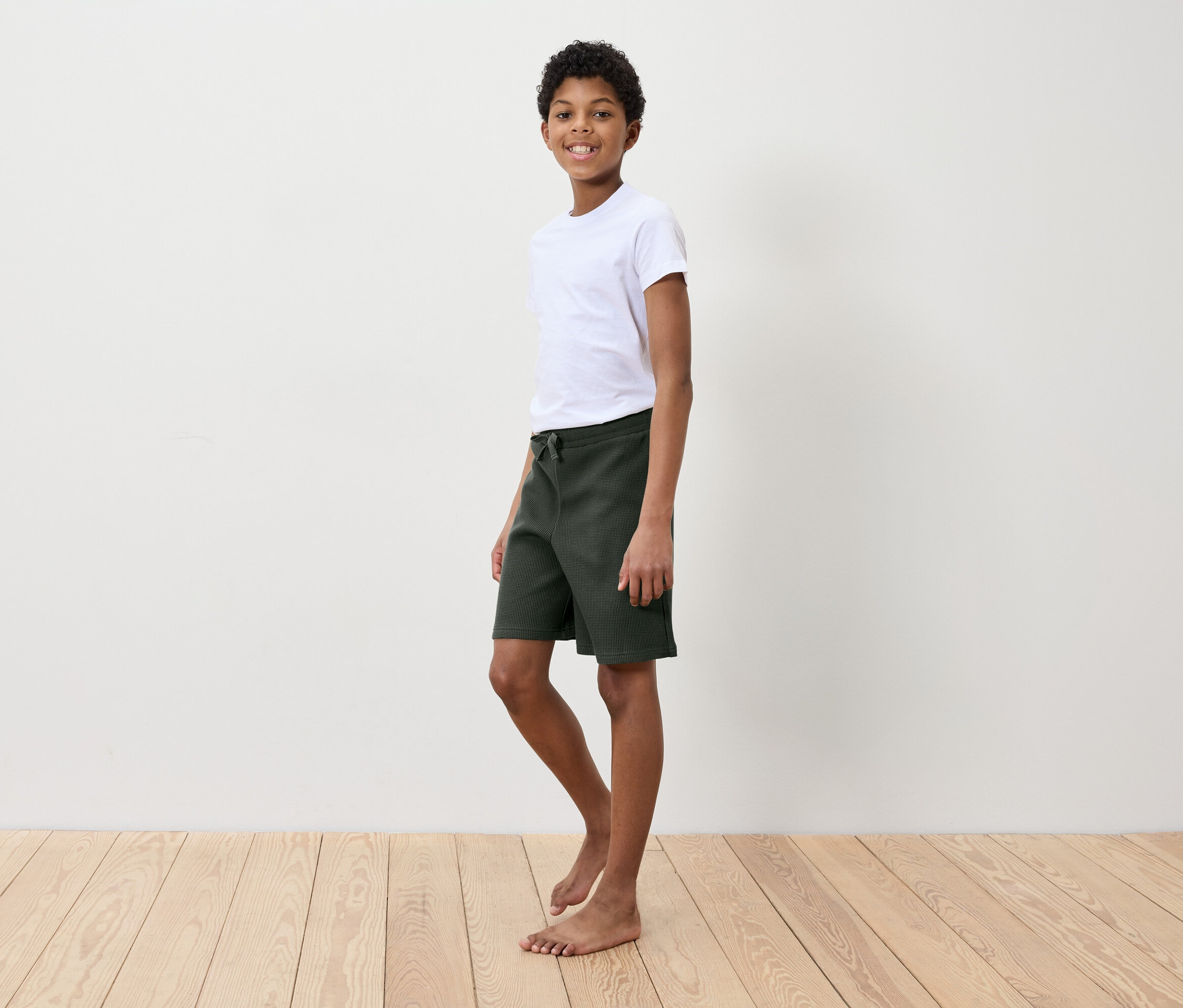 Chłopiec w białej koszulce i zielonych szortach stoi na drewnianej podłodze przed białą ścianą.