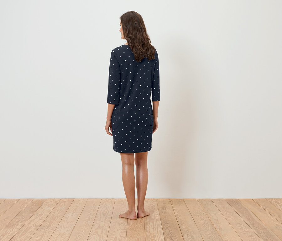 Kobieta stoi tyłem do kamery, ubrana w granatową sukienkę w białe kropki.