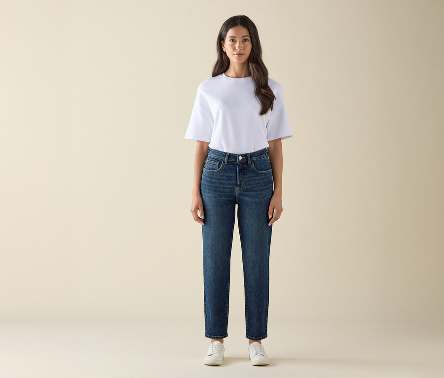 Kobieta stoi i ma na sobie spodnie dżinsowe typu mom – fason „Nyla” dark blue, biały T-shirt i białe tenisówki.