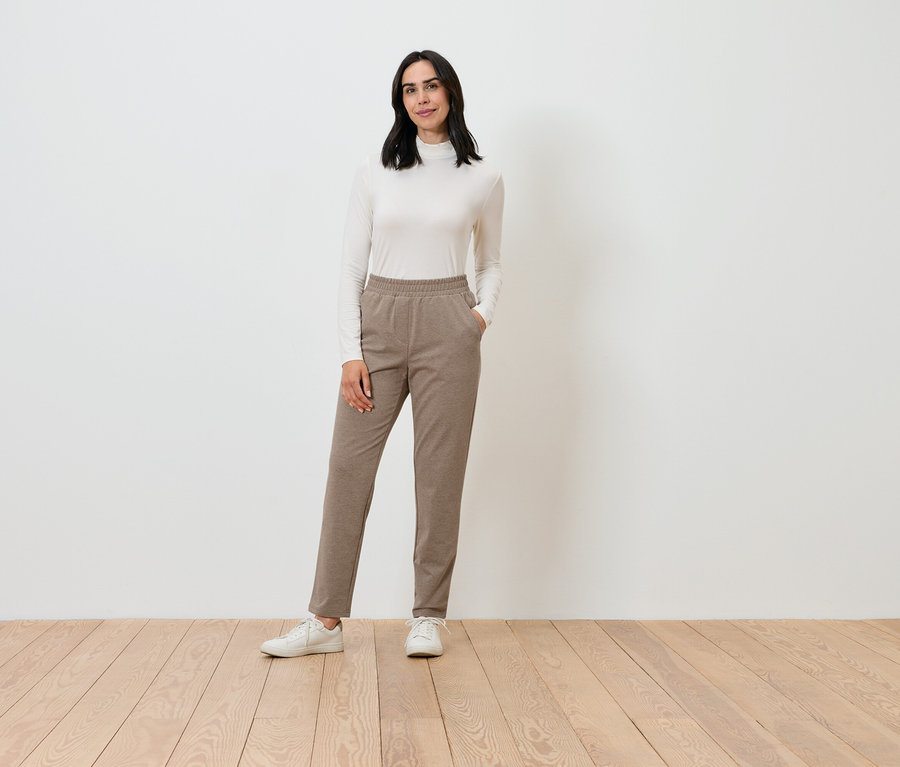 Kobieta pozuje w kremowym golfie i brązowych spodniach.