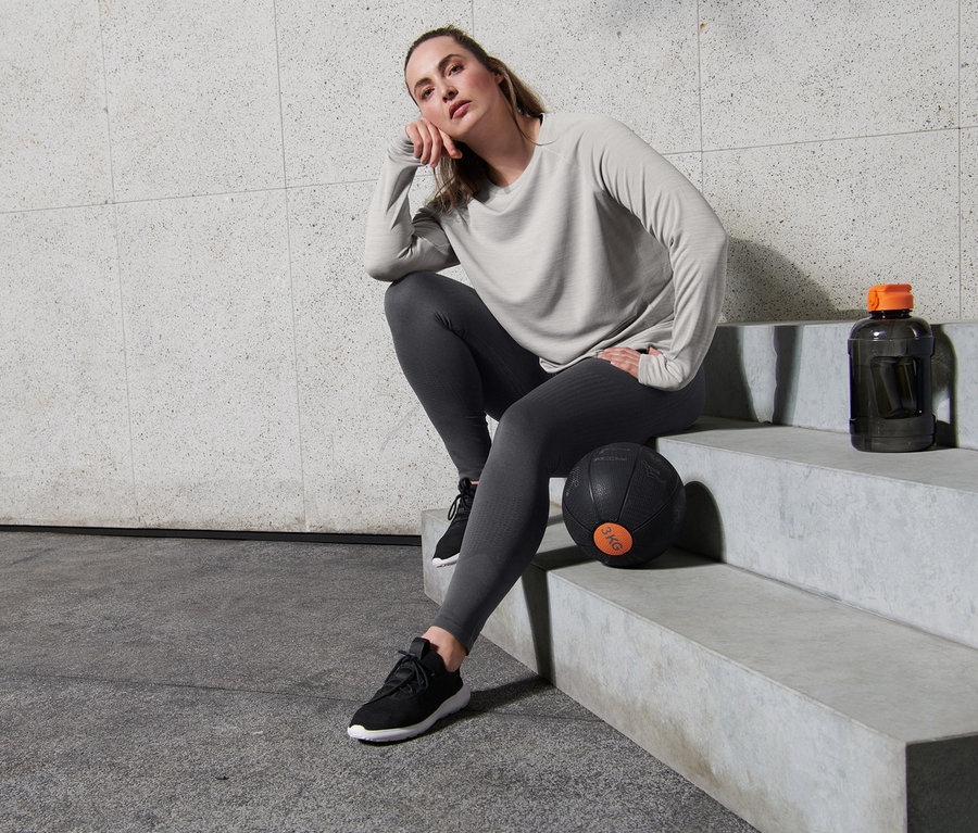 Kobieta siedzi na schodach w szarej bluzie funkcyjnej z długim rękawem i bezszwowych legginsach sportowych.
