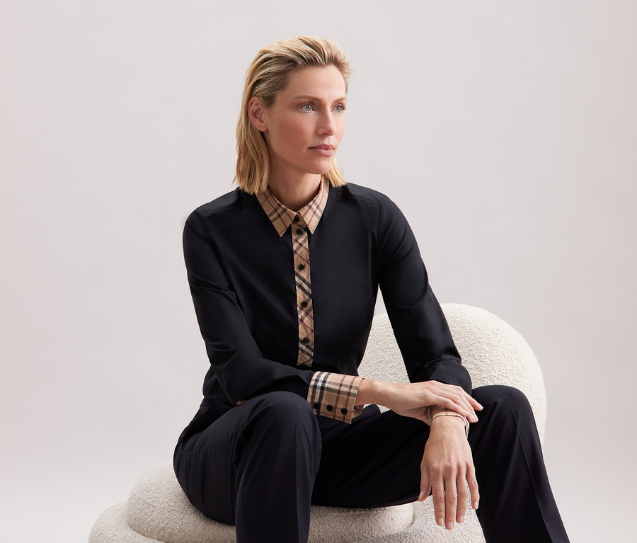 Blondwłosa kobieta siedzi na białym krześle, ubrana w czarne ubranie z detalami w kratę.