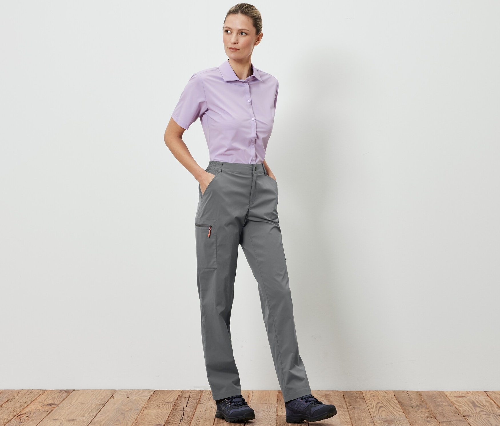 Kobieta stoi i pozuje w szarych spodniach outdoorowych z możliwością spakowania i jasno fioletowej koszuli.