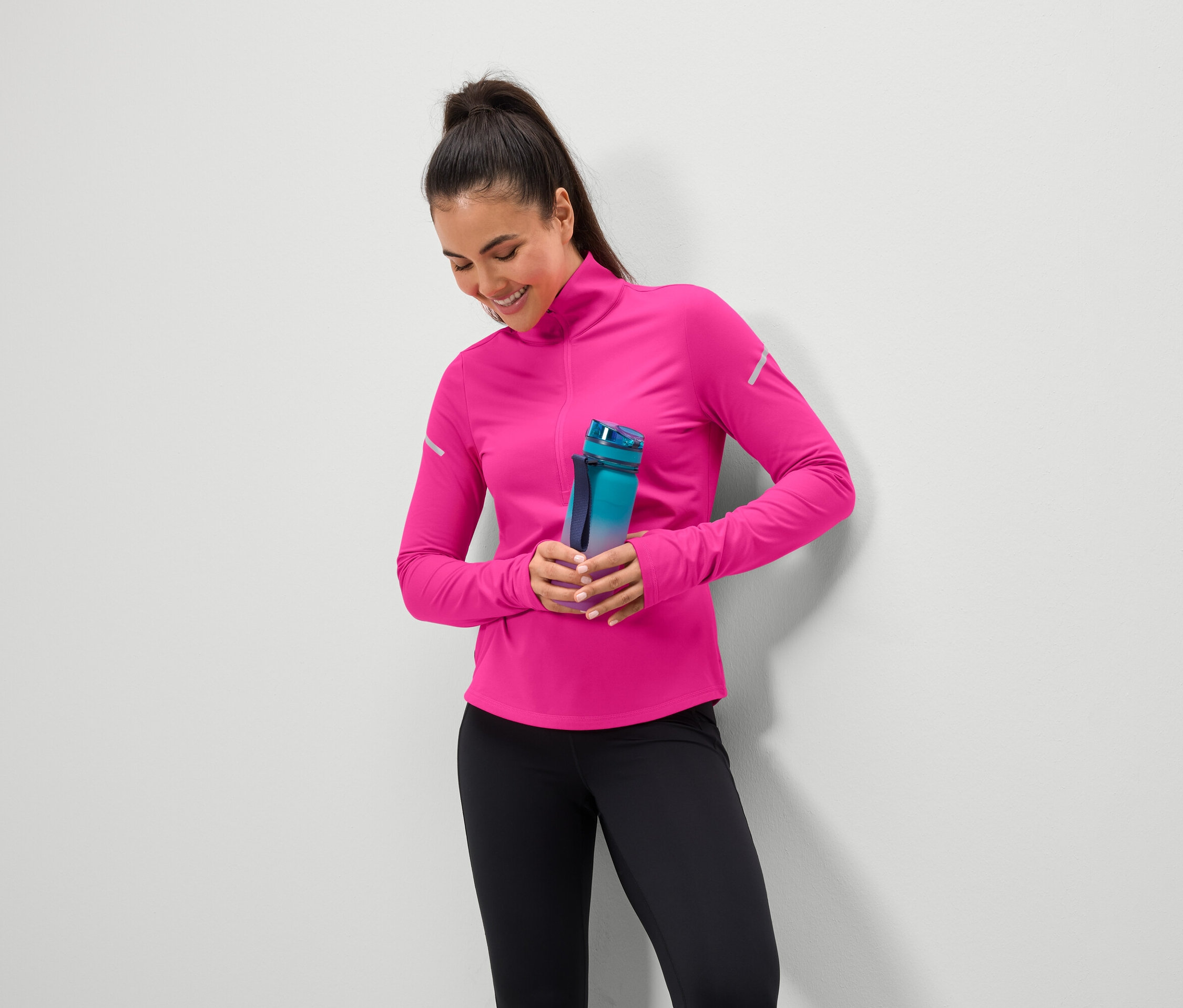 Uśmiechnięta kobieta trzyma niebiesko-zieloną butelkę na wodę, ma na sobie różowy top i czarne legginsy.