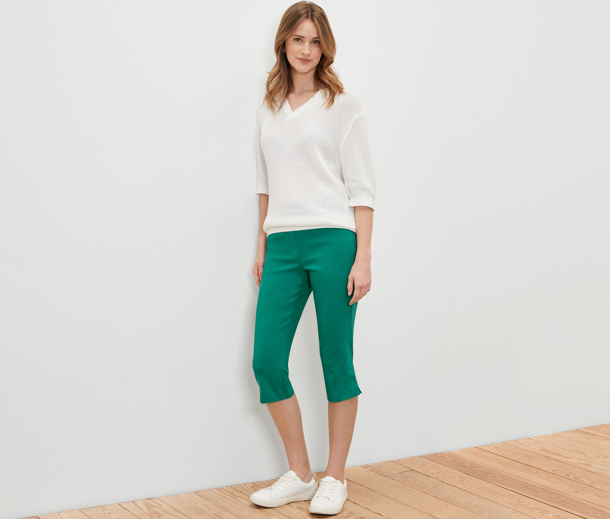 Kobieta pozuje w zielonych spodniach z bengaliny 3/4, białym swetrze i białych butach.