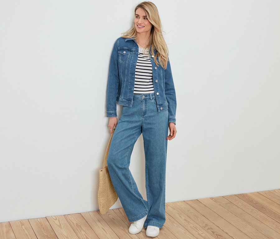 Blondynka opiera się o białą ścianę, ma na sobie kurtkę dżinsową, sweter w paski, dżinsy palazzo i trzyma torbę shopperkę z plecionki.