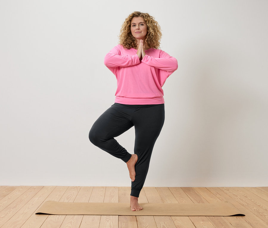 Kobieta o kręconych włosach ćwiczy jogę na beżowej macie, ubrana w różową bluzę i szare spodnie.