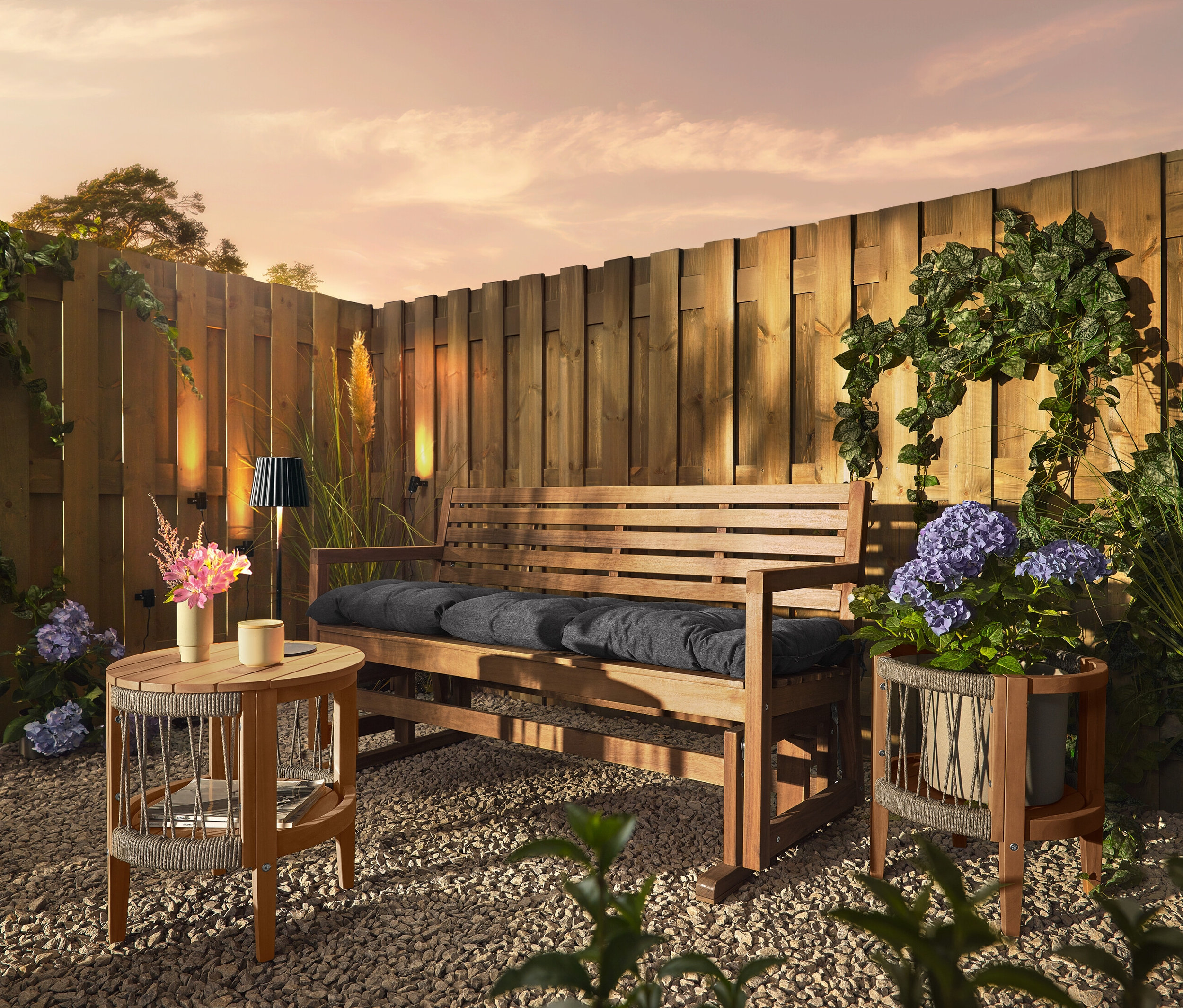 Drewniana ławka z poduszkami stoi na żwirowym podłożu przed drewnianym płotem. Na płocie zamontowane są 3 lampy solarne na rynnę dachową.