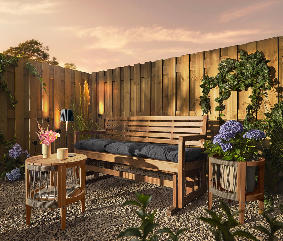 Drewniana ławka z poduszkami stoi na żwirowym podłożu przed drewnianym płotem. Na płocie zamontowane są 3 lampy solarne na rynnę dachową.