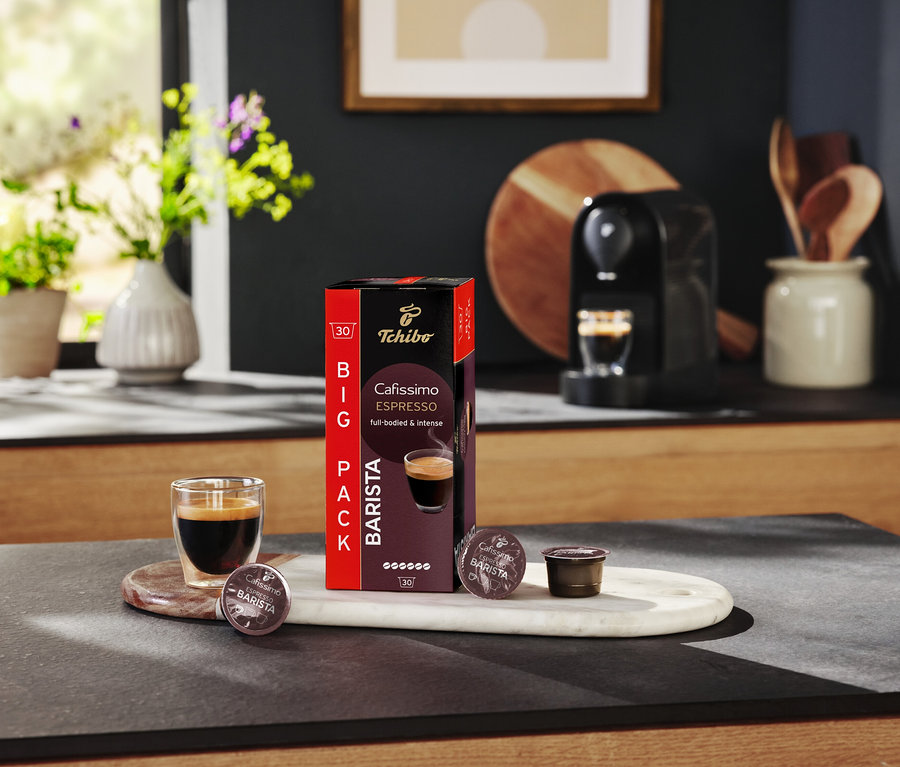 Szklanka espresso stoi obok opakowania kapsułek Tchibo Cafissimo Barista na stole kuchennym.