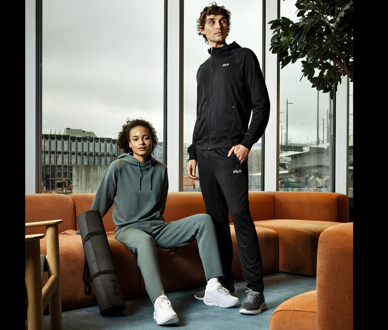 Młoda kobieta siedzi na pomarańczowej sofie, trzymając czarną matę do jogi. Obok stoi młody mężczyzna w czarnym dresie.