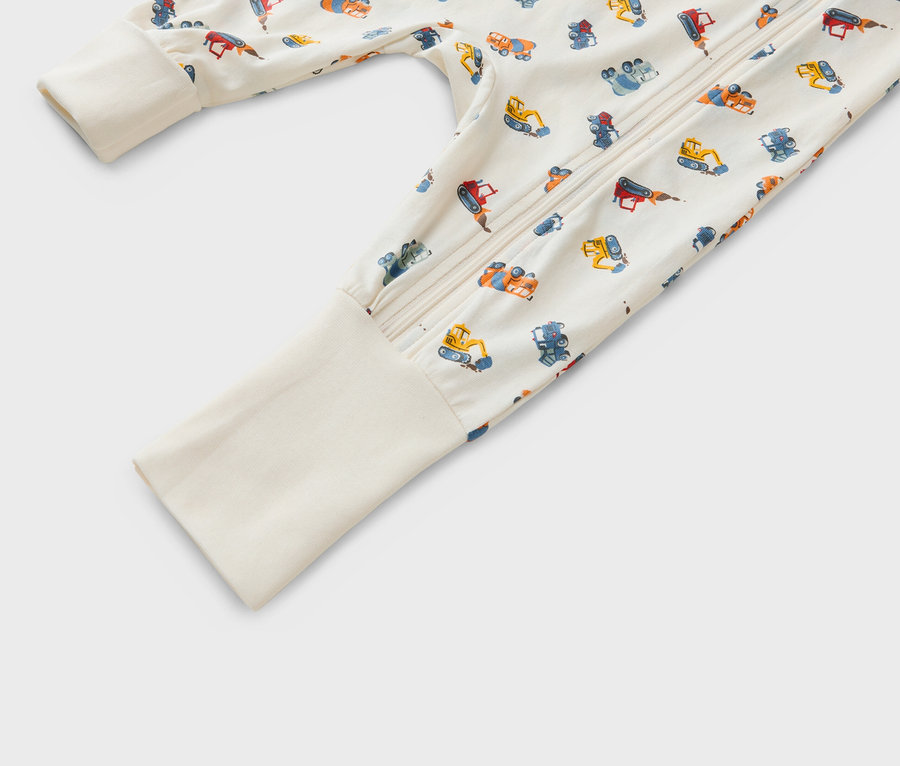 Zbliżenie na śpioszki niemowlęce zapinane na zamek, z wzorem przedstawiającym różne pojazdy budowlane na beżowym tle.