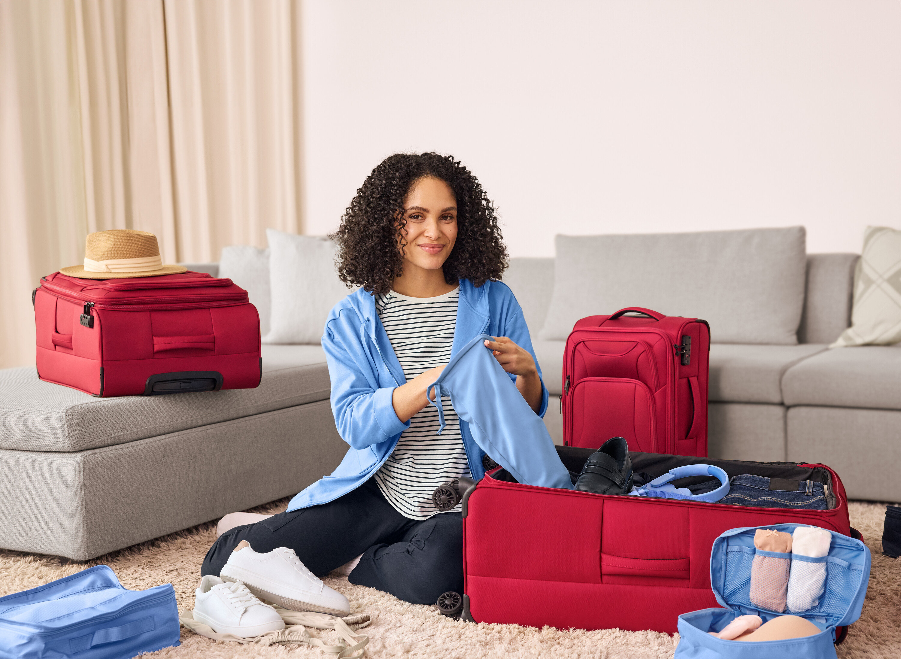 Kobieta siedzi na podłodze i pakuje granatowe spodnie Joggpants z kieszeniami cargo do czerwonej walizki. Obok niej leżą skórzane sneakersy.