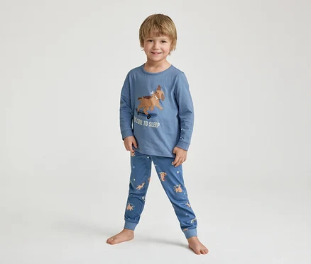 Uśmiechnięty mały chłopiec stoi boso w niebieskiej piżamie z nadrukiem psa na deskorolce i napisem "TOO COOL TO SLEEP".
