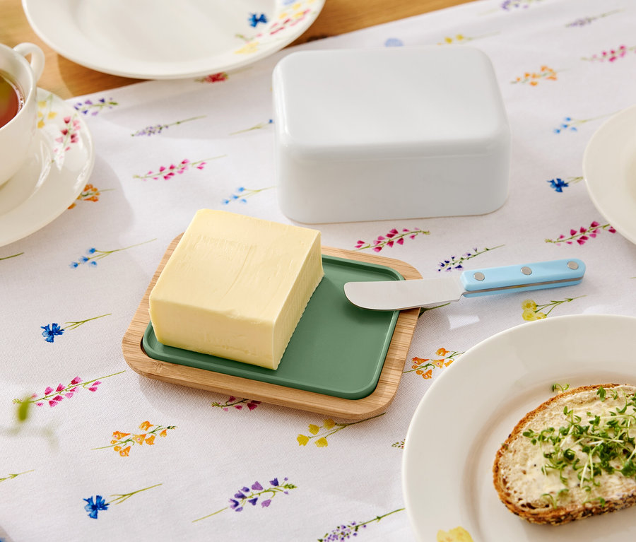 Na kwiecistym stole leży masło na drewnianym talerzu z zieloną wkładką, nóż do masła, maselniczka, filiżanka herbaty i talerz z kanapką z masłem.