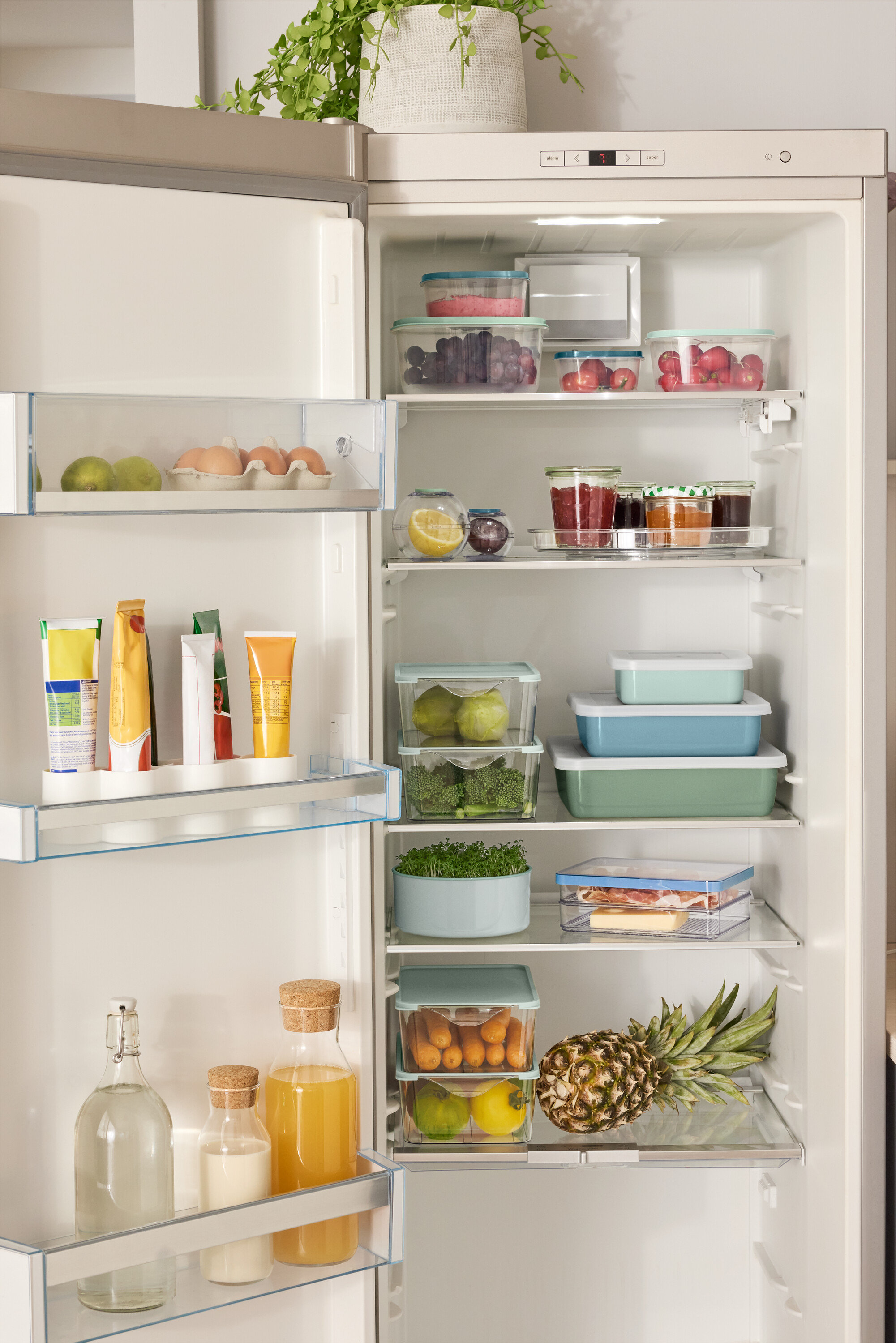 Otwarta lodówka wypełniona różnymi produktami spożywczymi, w tym 4 pudełka do przechowywania, 2 pojemniki spożywcze w kształcie kuli, 3 emaliowane formy do zapiekania z pokrywką i obracany talerz.