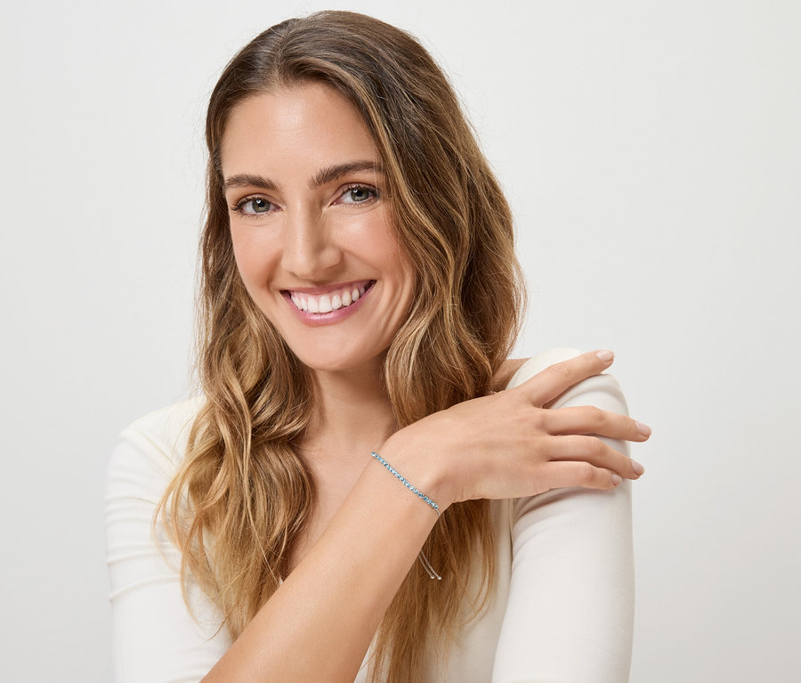 Uśmiechnięta kobieta z blond, falowanymi włosami i niebieską bransoletką na lewym nadgarstku na białym tle.