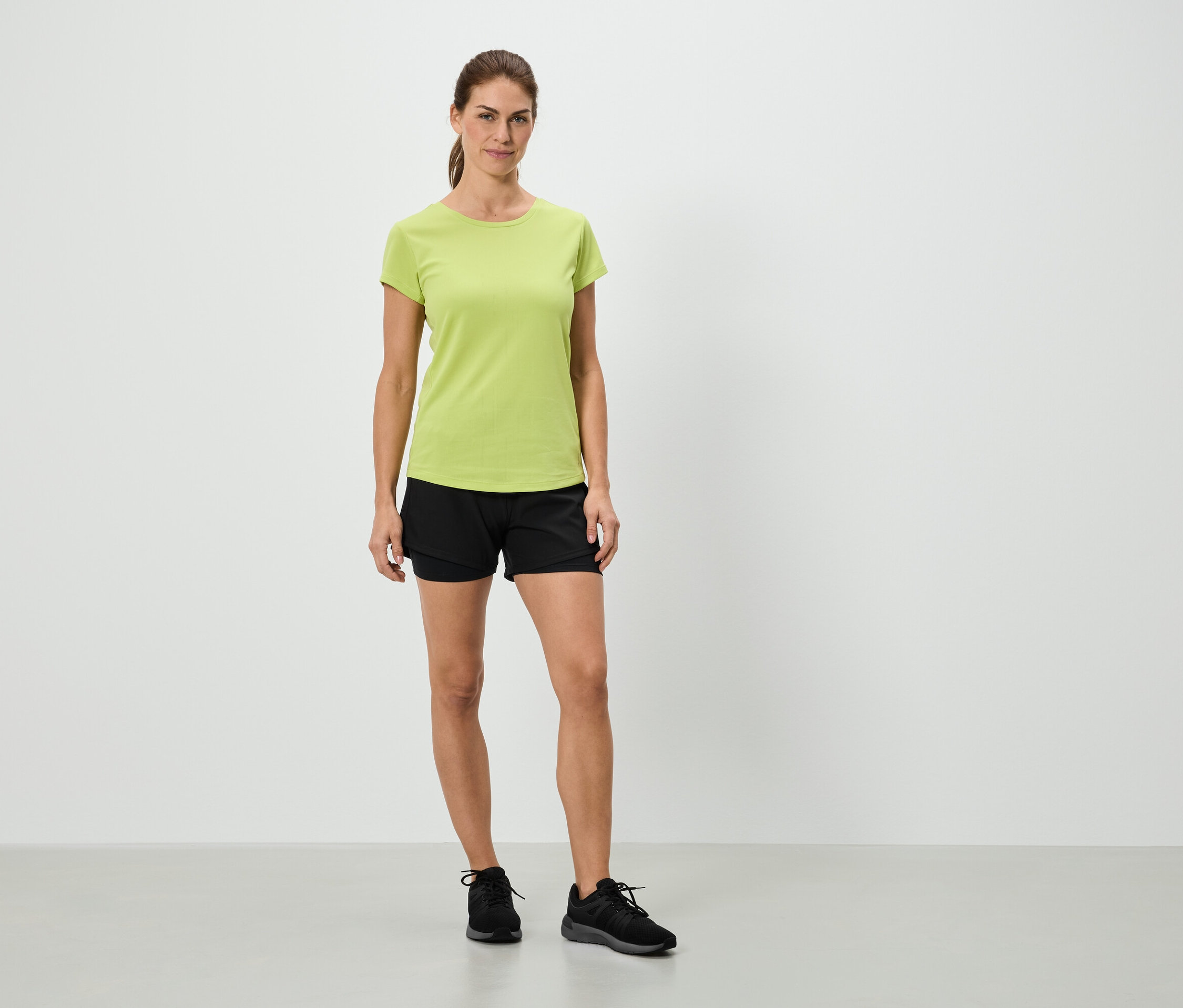 Kobieta stoi w jasnozielonej koszulce, czarnych szortach i czarnych butach sportowych na tle białej ściany.