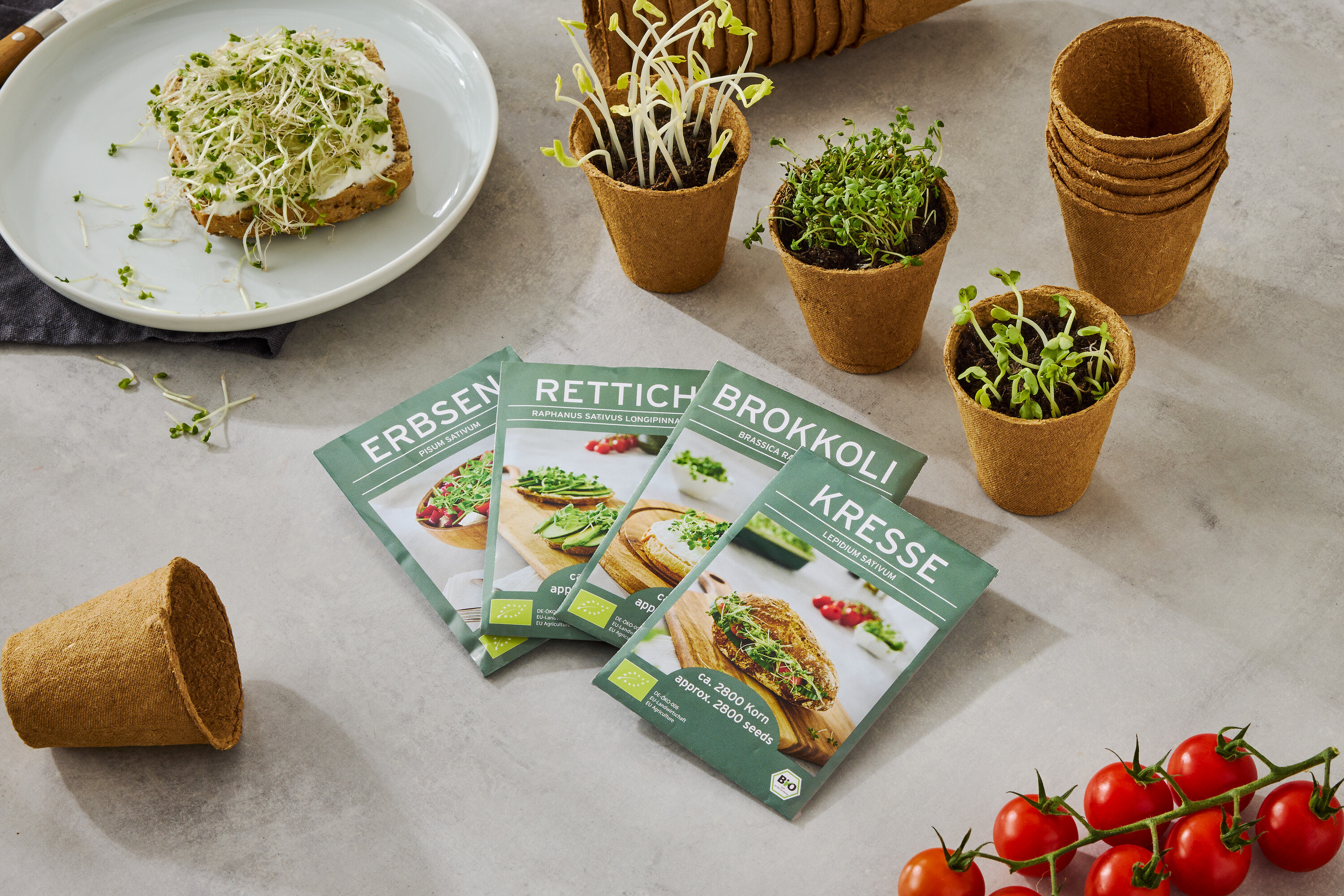 Talerz z chlebem posypanym kiełkami, wraz z czterema pudełkami nasion (grochu, rzodkiewki, brokułów, rzeżuchy), małymi doniczkami z rosnącymi kiełkami i gałązką pomidorów.