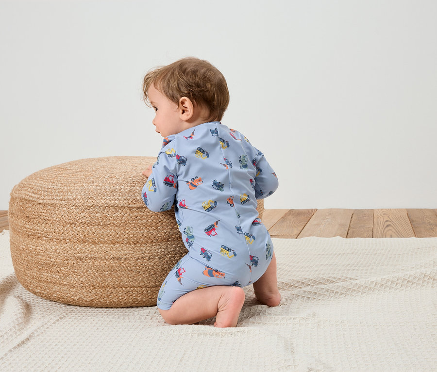 Dziecko w jasnoniebieskim kombinezonie we wzory z pojazdami budowlanymi klęczy na białym kocu obok okrągłego, plecionego pufa.