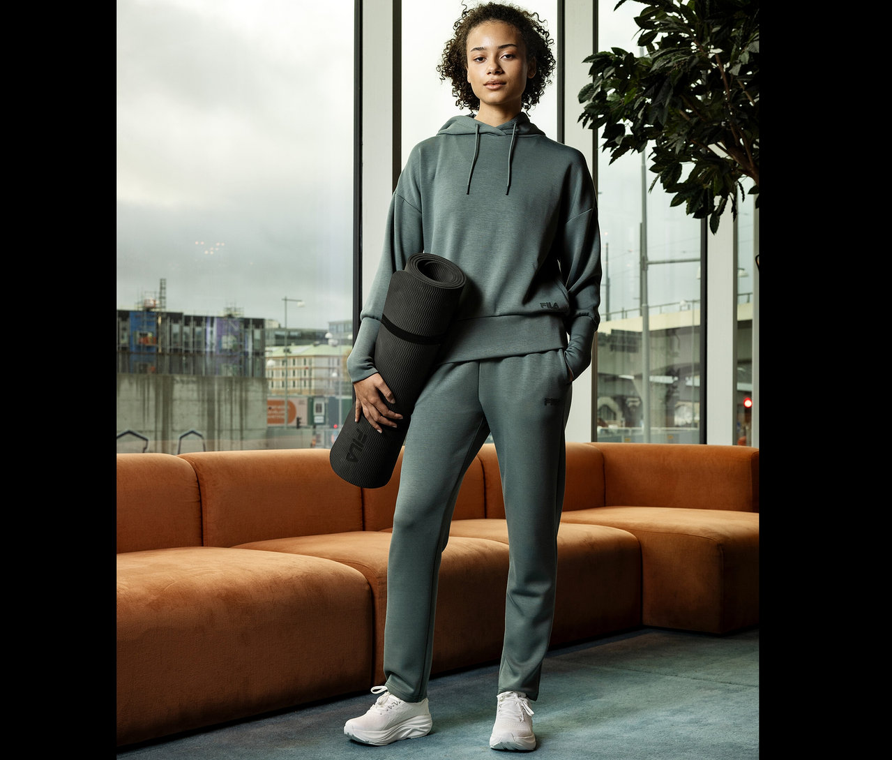 Kobieta w zielonym dresie trzyma matę do jogi na tle okna.