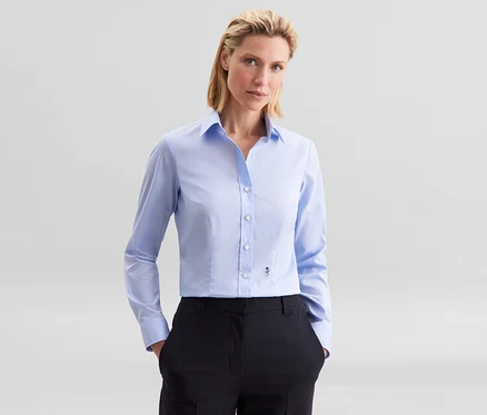 Kobieta w jasnoniebieskiej bluzce Seidensticker Slim Fit non iron i czarnych spodniach, z rękami w kieszeniach.
