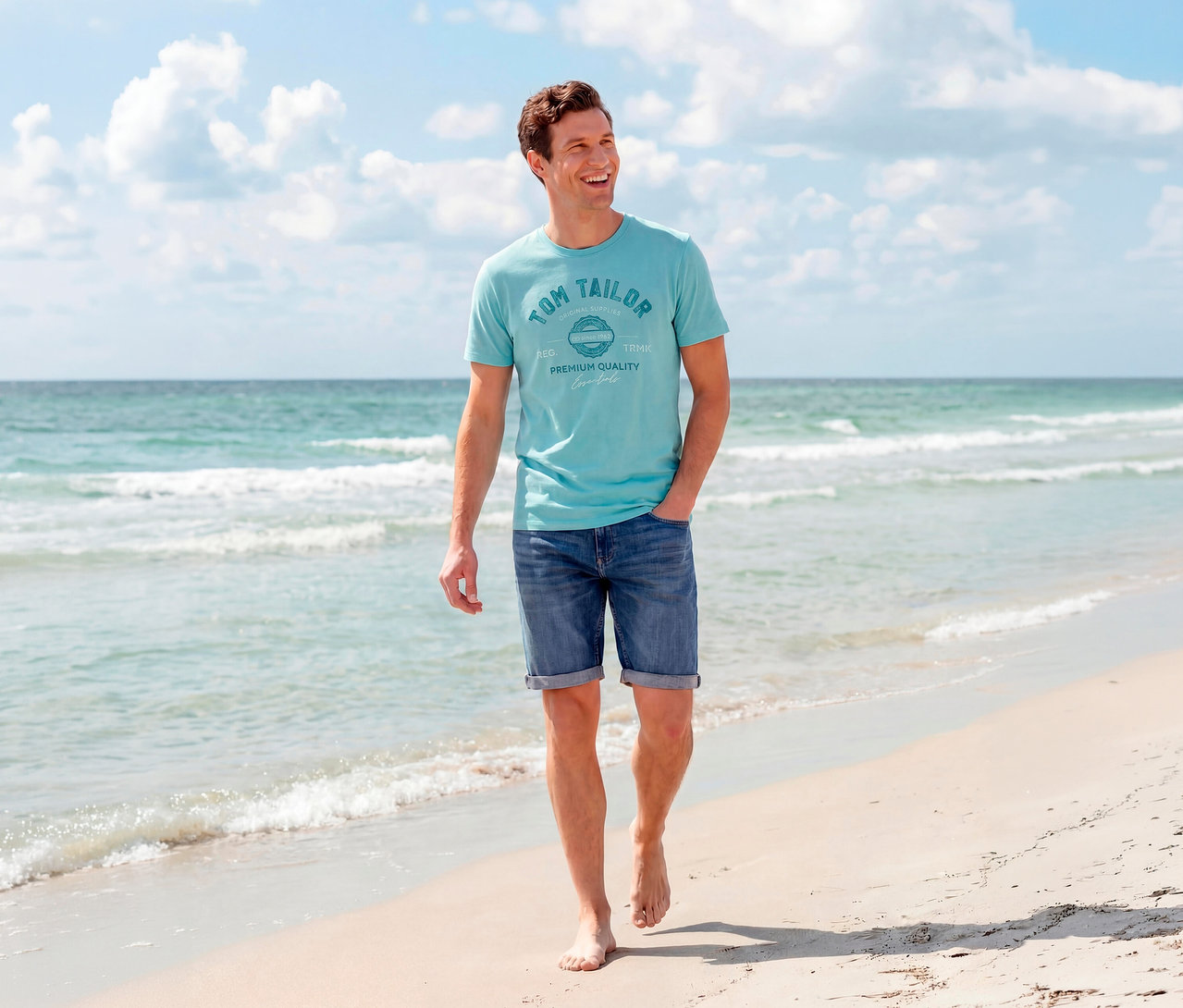 Uśmiechnięty mężczyzna idzie boso po piaszczystej plaży, ubrany w turkusową koszulkę z logo Tom Tailor i szorty dżinsowe Tom Tailor TTJOSH REGULAR SLIM w kolorze mid stone bright blue denim, z oceanem i niebem w tle.