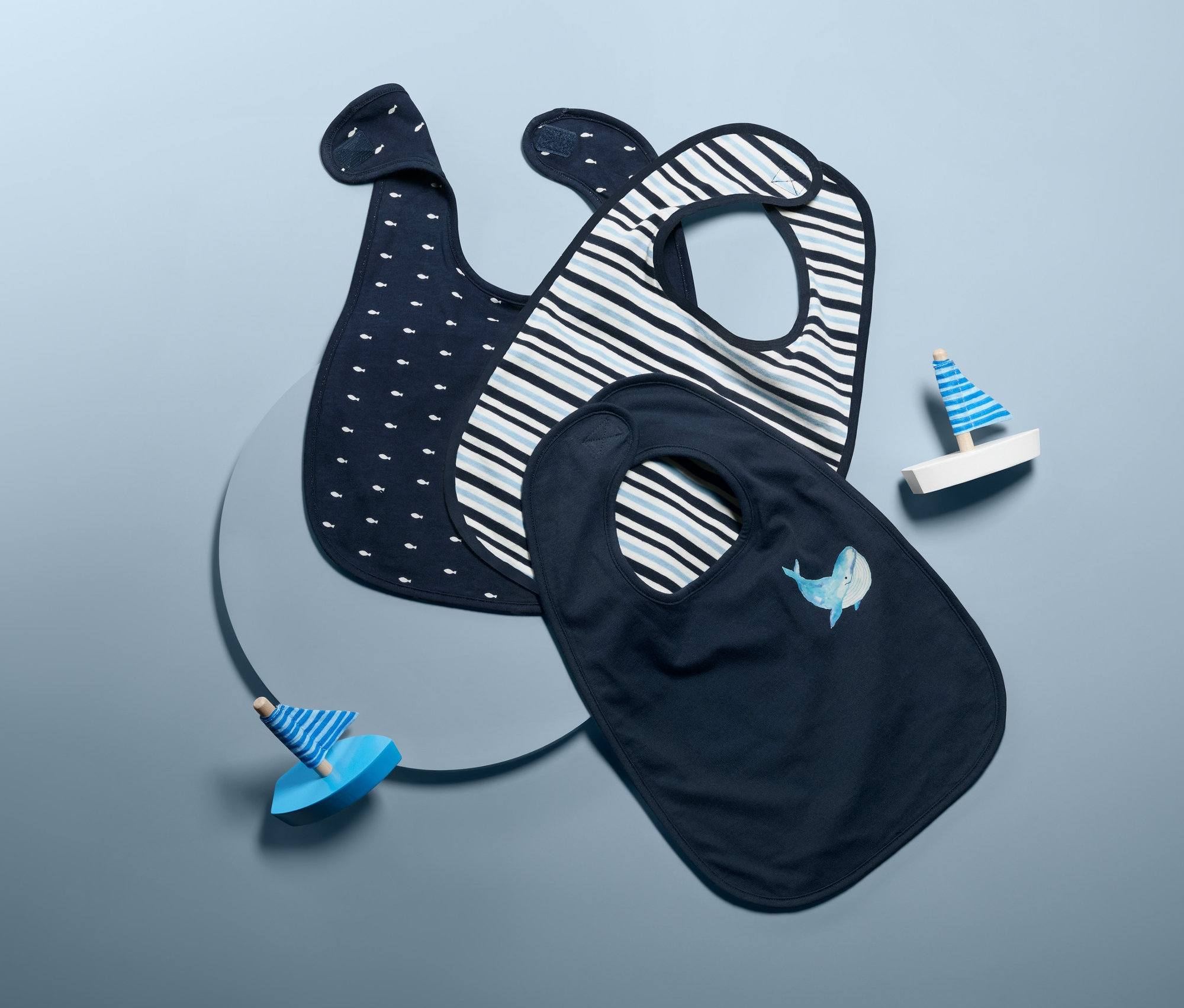 Trzy bawełniane śliniaki z nadrukiem i dwie drewniane zabawki na niebieskim tle.