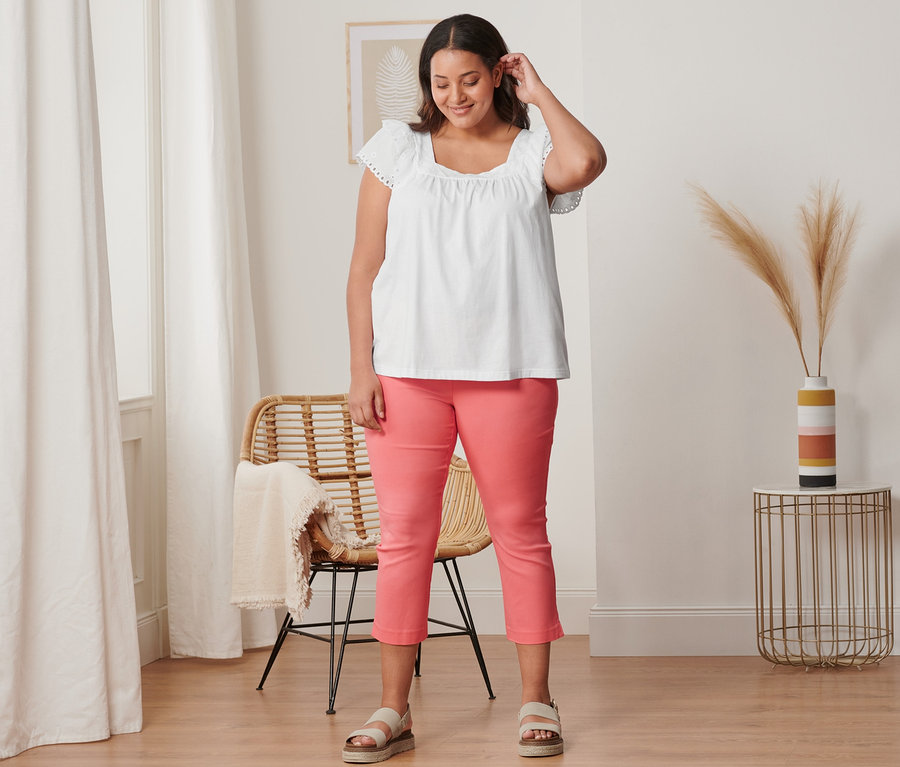 Kobieta ma na sobie koralowe spodnie stretchowe o długości 7/8, białą bluzkę z haftem i skórzane sandały na platformie.
