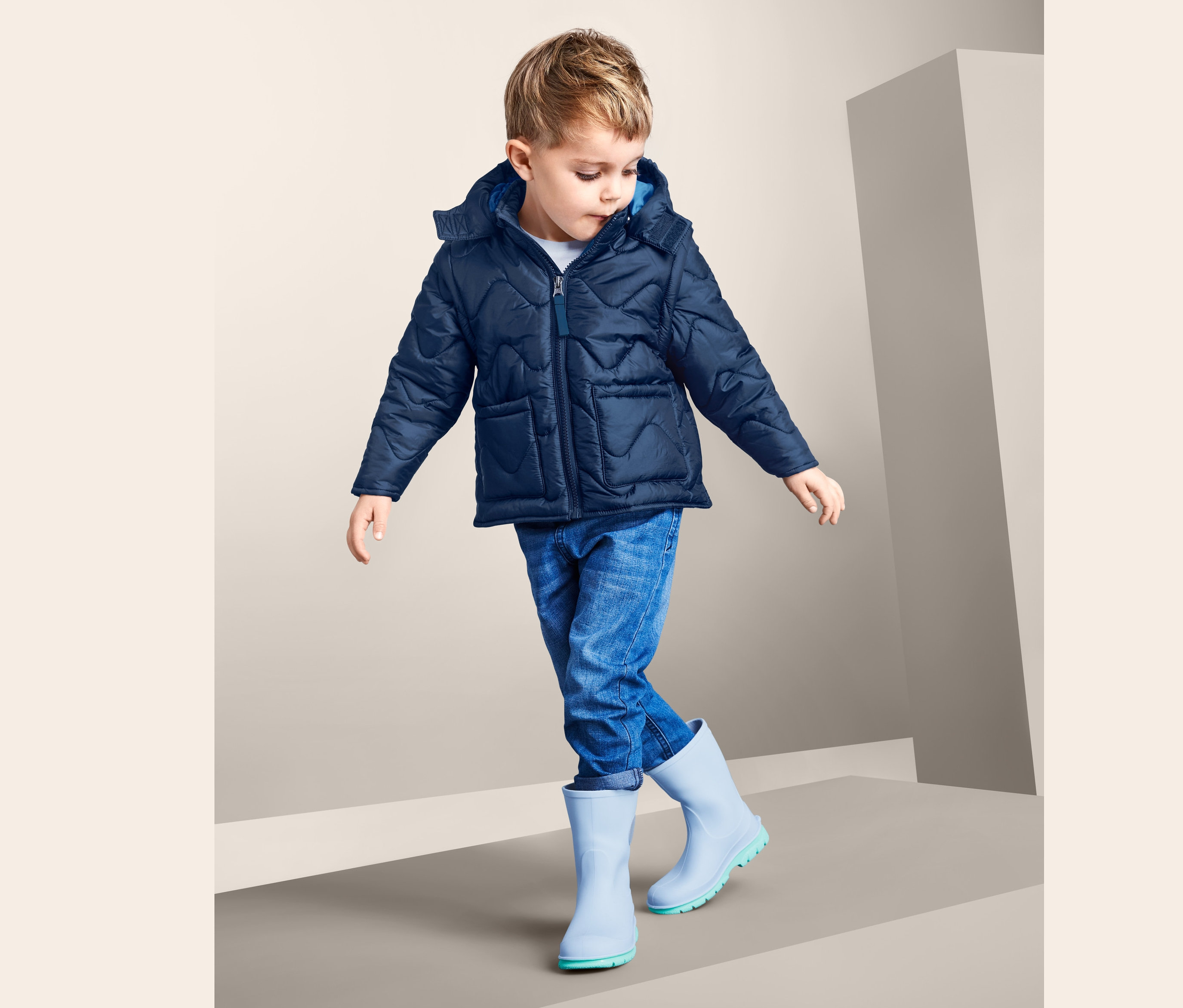 Chłopiec w niebieskiej kurtce pikowanej z odpinanymi rękawami, dżinsach i niebieskich kaloszach stoi na szarej powierzchni.