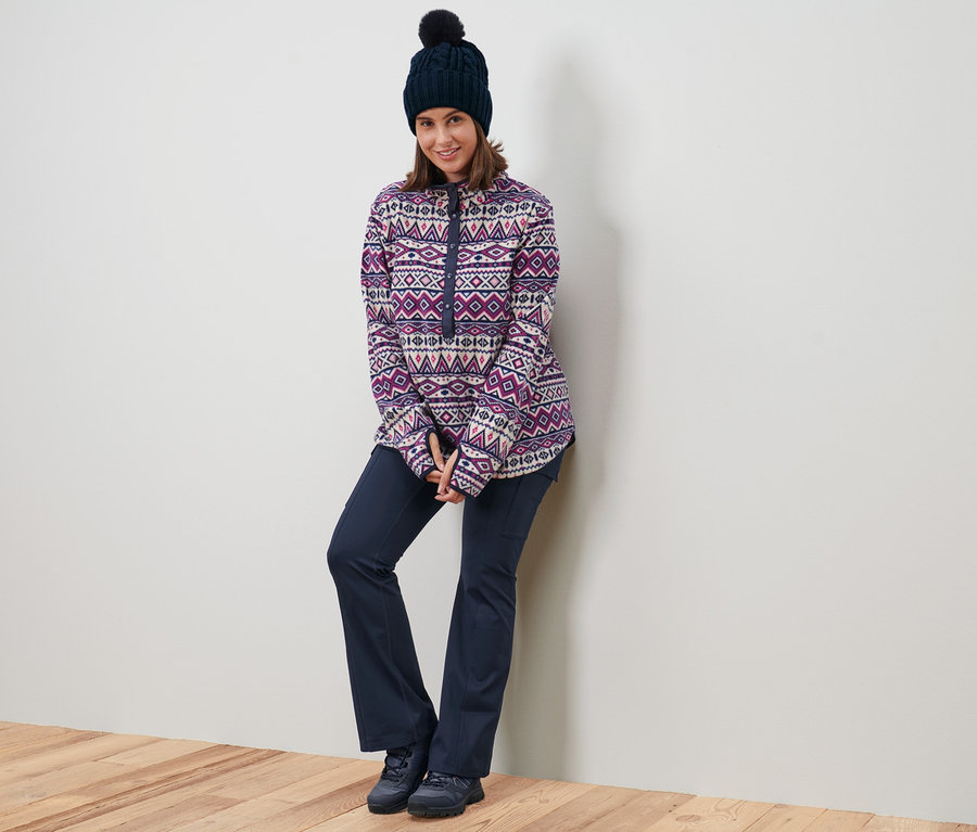 Kobieta oparta o ścianę ma na sobie bluzę z polaru, czapkę z dzianiny i termoaktywne spodnie.