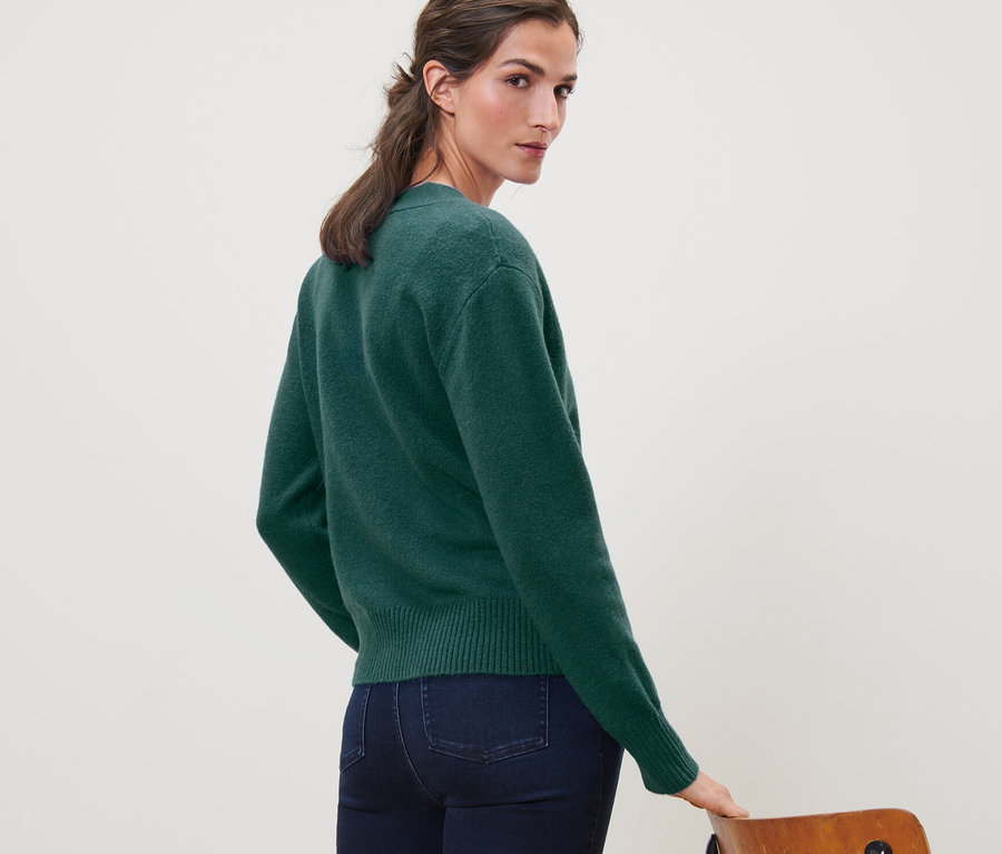 Kobieta spogląda przez ramię, ubrana w ciemnozielony rozpinany sweter z zawartością wełny i dżinsy.
