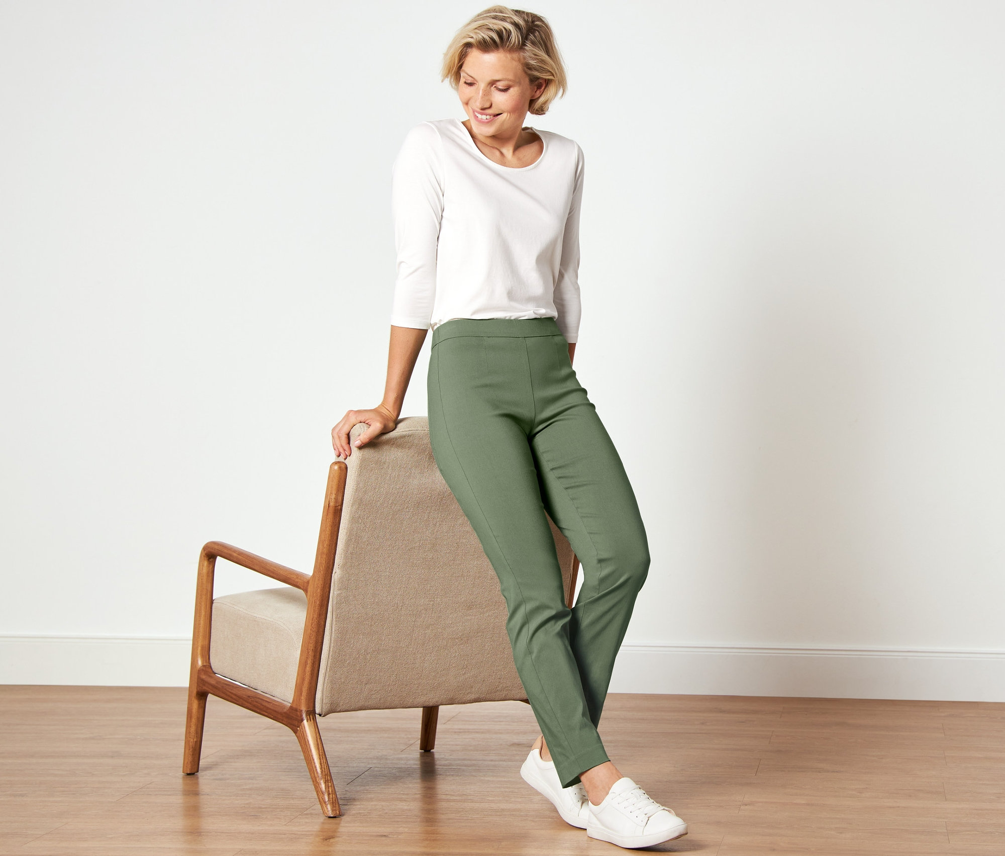 Kobieta opiera się o krzesło, ubrana w białą koszulę i spodnie stretchowe.